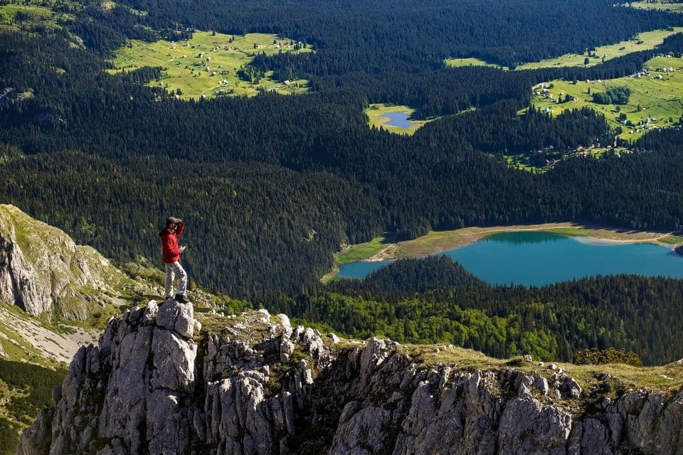 Una persona con chaqueta roja se alza en una cumbre rocosa, contemplando un valle con dos lagos azules y densos bosques.