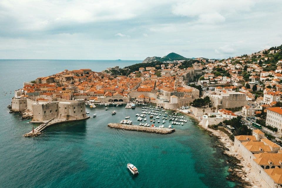 Aerial view of a historic walled city with orange-tiled roofs on a peninsula, with boats in a small harbor.