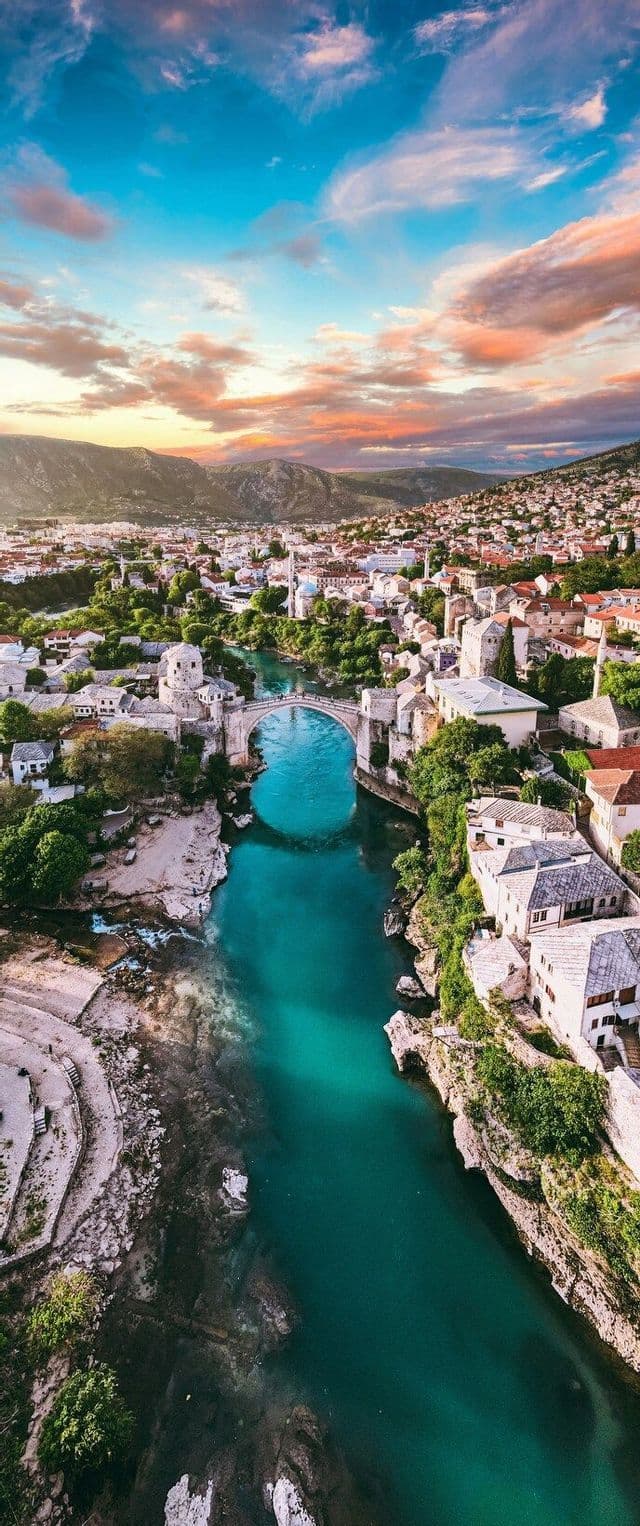 Un ponte ad arco in pietra attraversa un fiume turchese che scorre attraverso una città storica al tramonto, con montagne in lontananza.