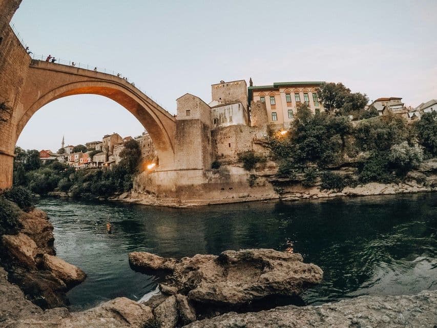 Un gran puente de arco de piedra cruza un río al atardecer, con edificios históricos y árboles bordeando la orilla rocosa del río.