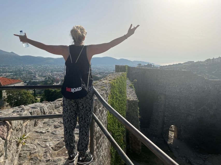 A woman with a WeRoad backpack stands on a stone wall with her arms outstretched, overlooking a coastal city from a high vantage point.