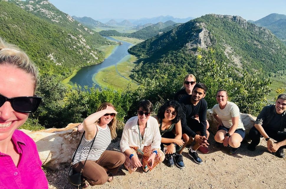 Un gruppo WeRoad in viaggio posa per una foto da un punto panoramico che domina un fiume tortuoso e una valle con verdi colline.