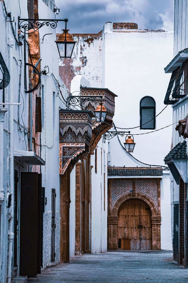 A narrow street with whitewashed buildings, ornate lanterns, and detailed tilework leading to a large arched wooden door.