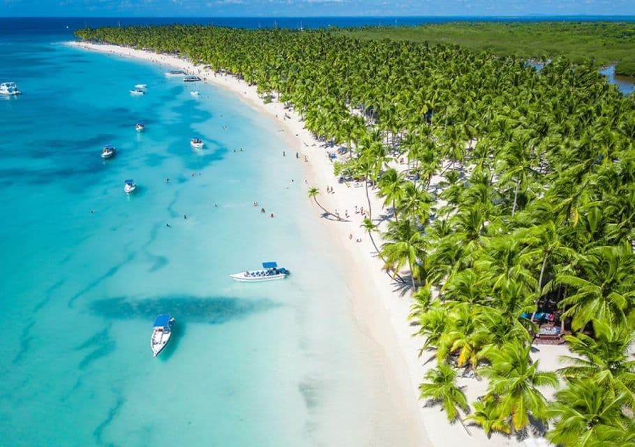 Luftaufnahme eines tropischen weißen Sandstrandes, gesäumt von Palmen, mit Booten im klaren türkisfarbenen Wasser und Menschen, die baden.