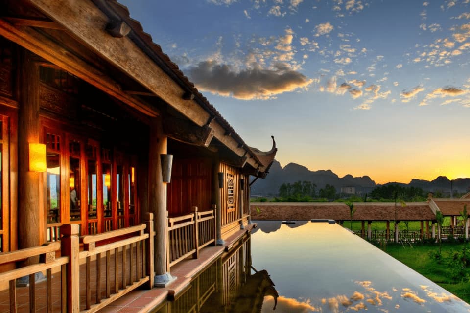 Ein traditionelles Holzgebäude neben einem Infinity-Pool, der den bewölkten Himmel bei Sonnenuntergang widerspiegelt, mit fernen Bergen im Blick.
