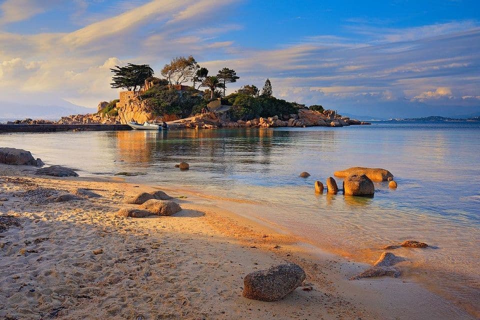 Una vista da una spiaggia sabbiosa attraverso acque calme verso un piccolo isolotto roccioso con alberi, sotto un cielo blu e nuvoloso.