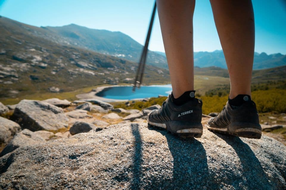 Una persona con scarpe da trekking sta su una grande roccia con un bastone da trekking, affacciata su un lago in una valle di montagna.