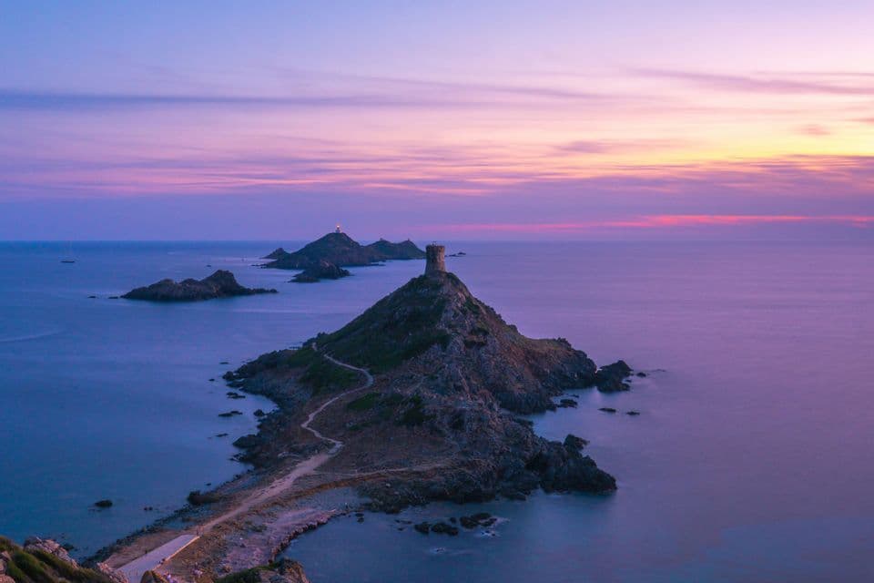 Una torre in pietra su una penisola rocciosa si protende in un mare calmo con un faro in lontananza sotto un cielo al tramonto viola.