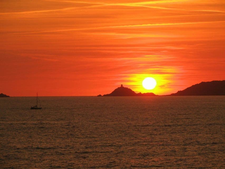 Il sole tramonta sull'oceano, stagliando un'isola con una torre e una barca a vela sull'acqua, tutto sotto un cielo arancione.
