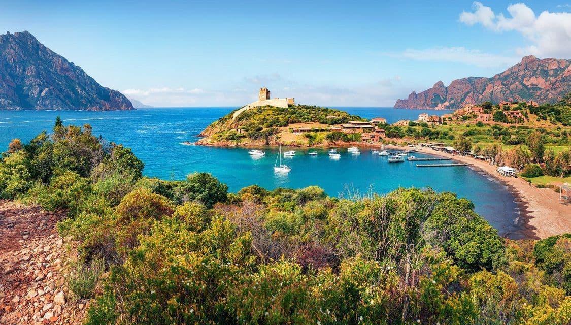 Vista dall'alto di una baia turchese con barche a vela ormeggiate, un villaggio costiero e una torre in pietra su un promontorio.