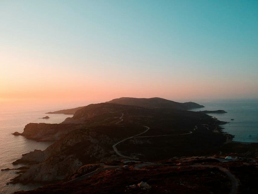 Una strada tortuosa si snoda lungo una penisola collinare che si protende nel mare, sotto un cielo al tramonto dai colori vivaci.