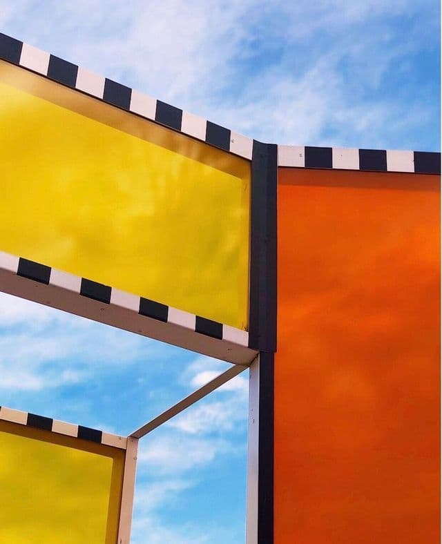 Una vista dal basso di una struttura moderna con pannelli traslucidi gialli e arancioni su un cielo blu con nuvole.