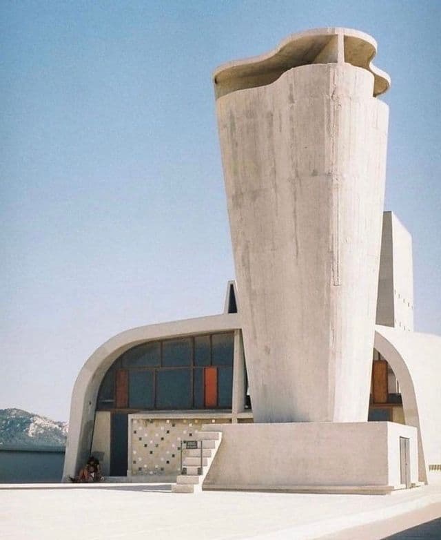 L'esterno di un edificio modernista in cemento con una torre prominente dalla forma unica e pareti curve, sotto un cielo azzurro chiaro.