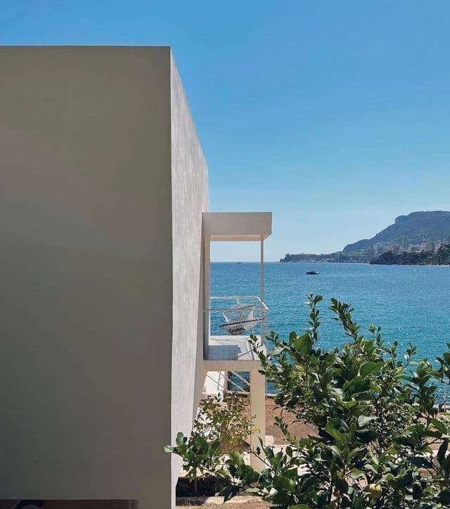 Un moderno edificio bianco con balconi si affaccia sul mare azzurro brillante, con una costa distante e vegetazione verde.
