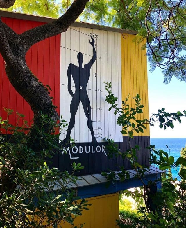 Il murale 'Modulor' di Le Corbusier su una facciata colorata di un edificio, incorniciato da alberi sullo sfondo del mare.