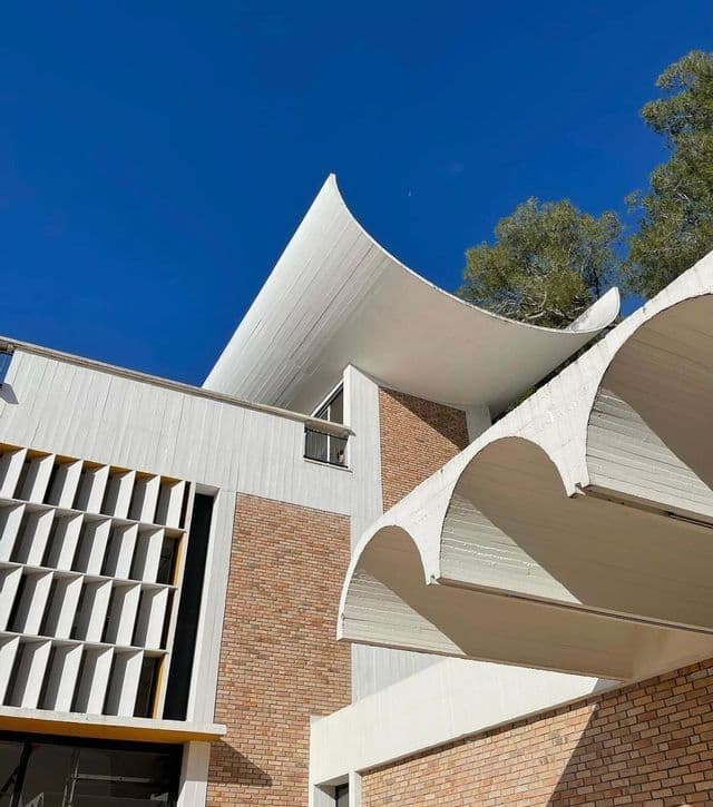 Un'inquadratura dal basso di un edificio moderno con un caratteristico tetto bianco curvo, muri in mattoni e una facciata a griglia contro un cielo azzurro intenso.