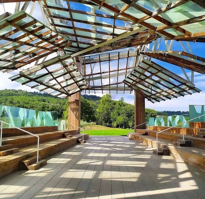Un padiglione all'aperto con tetto in legno e vetro getta ombre su sedute in legno a gradoni, con vista su un rigoglioso paesaggio verde.