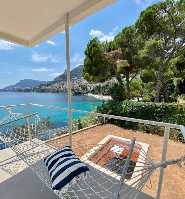 Un'amaca con un cuscino a righe bianche e blu su una terrazza con vista su una città costiera e una baia turchese.
