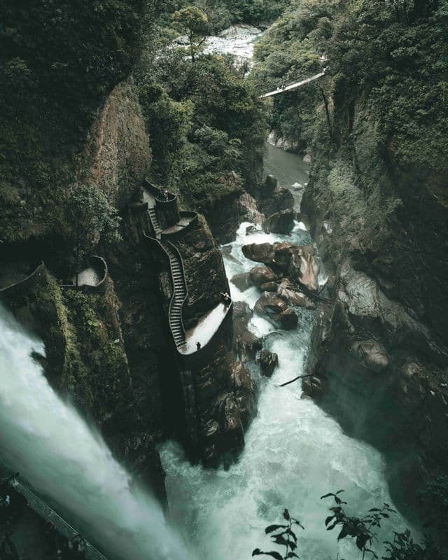 Una vista aérea de una sinuosa escalera de piedra que desciende por un acantilado junto a una potente cascada en un exuberante y verde desfiladero.
