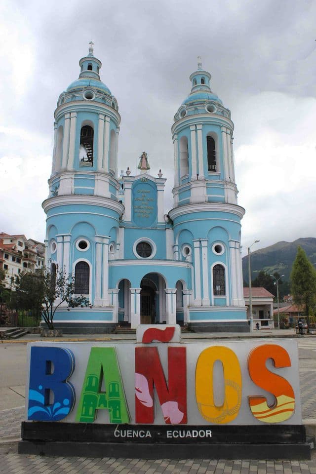 Un letrero colorido de 'Baños Cuenca - Ecuador' se alza en una plaza frente a una gran iglesia azul y blanca con torres gemelas.