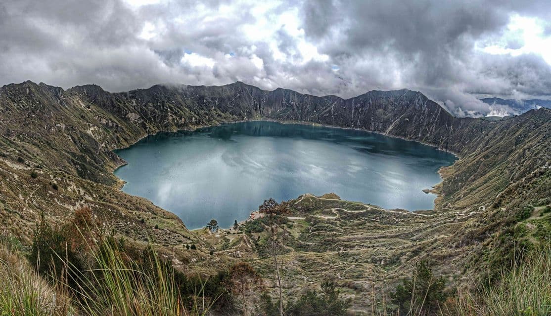 Una vista panorámica de un lago azul profundo dentro de un gran cráter de montaña bajo un cielo lleno de nubes grises y densas.