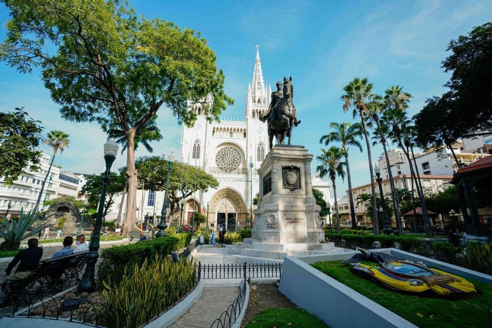 Una estatua ecuestre se alza en un parque de la ciudad frente a una gran catedral blanca, rodeada de palmeras y vegetación.