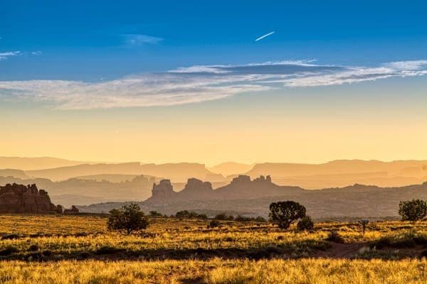 Un prato erboso con alberi sparsi si trova di fronte a strati di formazioni rocciose velate, sotto un cielo dorato e azzurro al tramonto.
