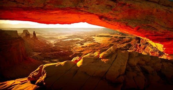 Un vasto paesaggio di canyon con formazioni rocciose, visto da sotto un grande arco di roccia rossa all'alba.