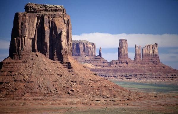 Imponenti butte e guglie di arenaria rossa si innalzano da un vasto paesaggio desertico sotto un cielo azzurro parzialmente nuvoloso.