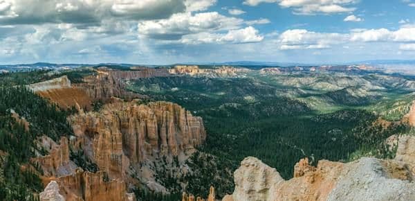 Vista panoramica di un vasto canyon con guglie di roccia arancioni, una fitta foresta verde e un cielo parzialmente nuvoloso.