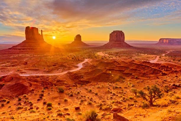 Il sole al tramonto proietta un bagliore dorato su un paesaggio desertico con grandi butte di arenaria e una strada tortuosa.