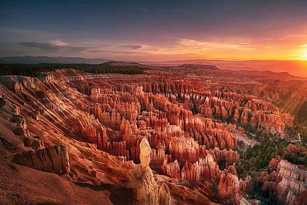 Un vasto canyon, pieno di camini delle fate e guglie di roccia rossa, illuminato dalla calda luce di un tramonto, con una foresta sullo sfondo.