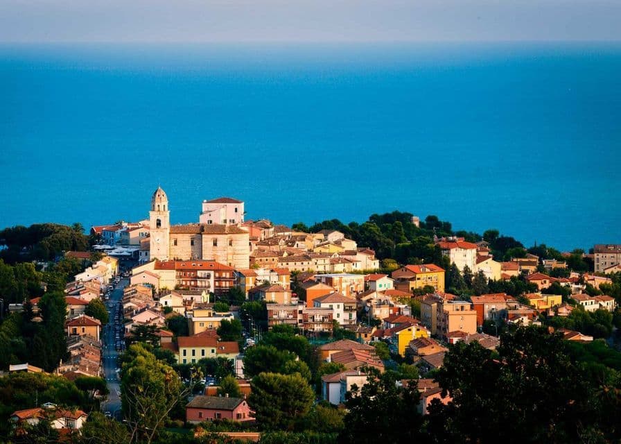 Un borgo costiero con edifici colorati e una chiesa si erge su una verde collina che domina il mare blu profondo.