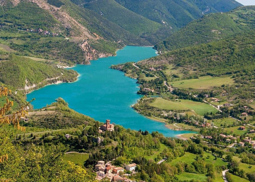 Una vista aerea di un vivace lago turchese che serpeggia in una lussureggiante valle verde, punteggiata da piccoli villaggi e un castello storico.