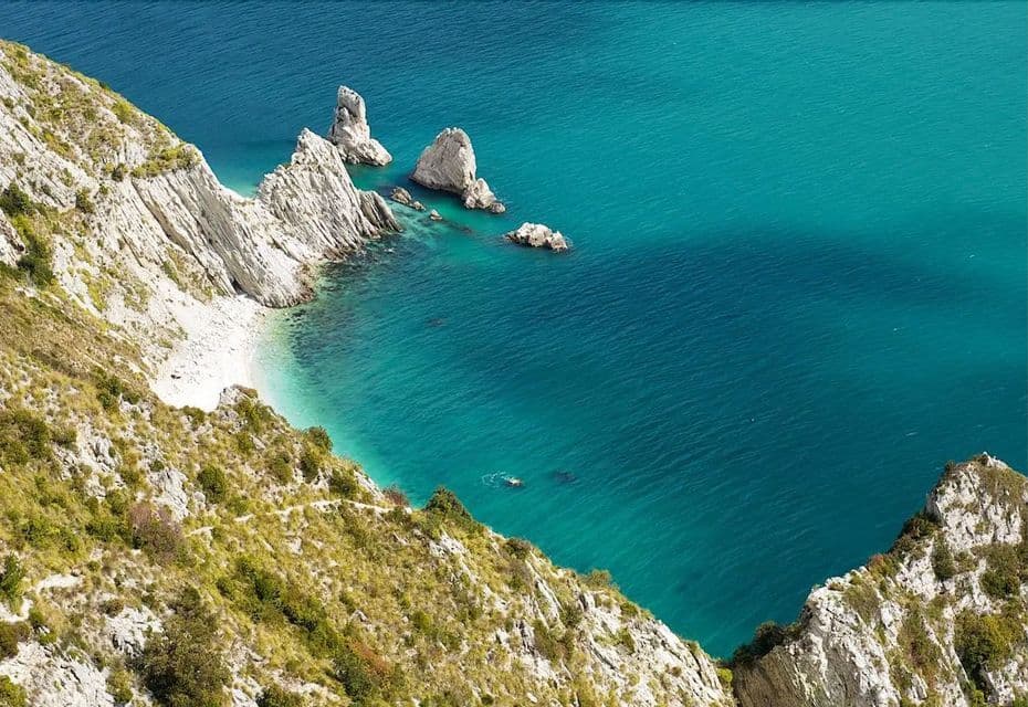Vista aerea di una persona che nuota in acqua turchese vicino a scogliere bianche e una spiaggia di ciottoli isolata.