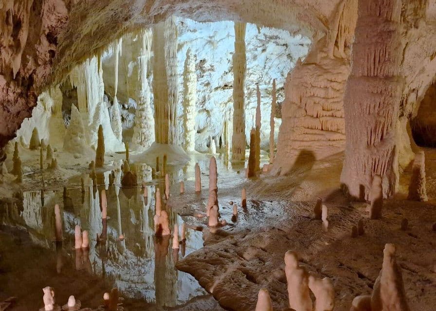 L'interno di una grotta illuminata con stalattiti e stalagmiti riflesse in uno specchio d'acqua immobile.