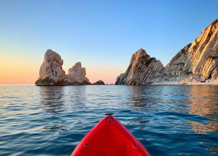 Vista dalla prua di un kayak rosso su acqua calma, di fronte a imponenti faraglioni e una costa rocciosa al tramonto.