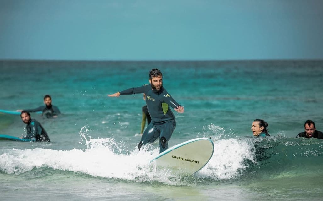 Un uomo in muta si bilancia su una tavola da surf, cavalcando un'onda durante un viaggio di gruppo WeRoad con altre persone nell'oceano.