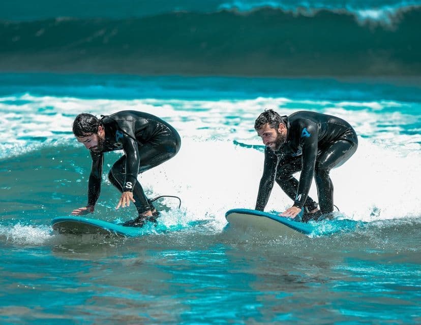 Due uomini in mute nere si accovacciano sulle loro tavole da surf, cavalcando una piccola onda in acqua turchese.