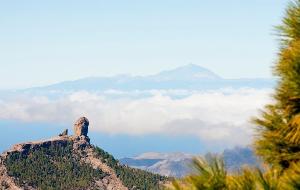 Un'imponente formazione rocciosa su una vetta montuosa boscosa si erge sopra un mare di nuvole, con un vulcano lontano all'orizzonte.