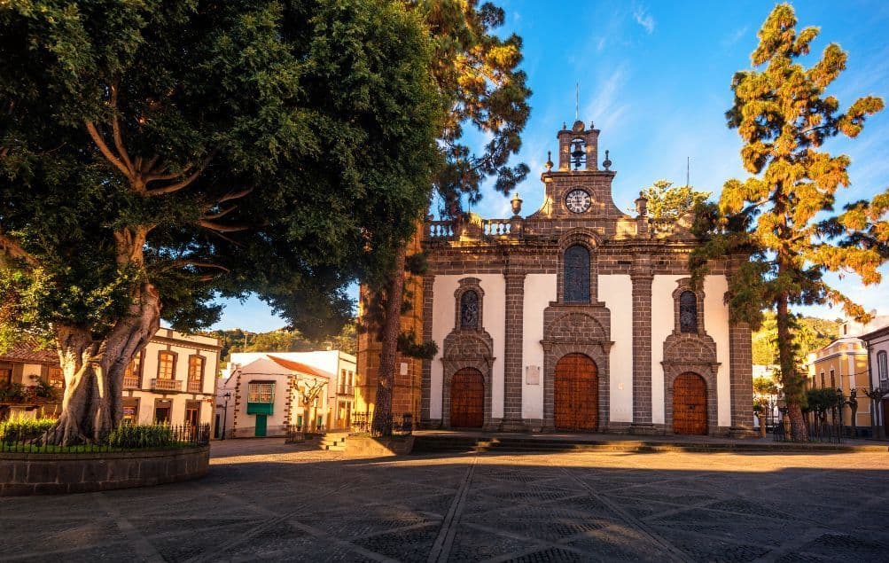 Una storica chiesa in pietra con un campanile si erge in una piazza lastricata, illuminata dalla luce dorata del cielo blu.