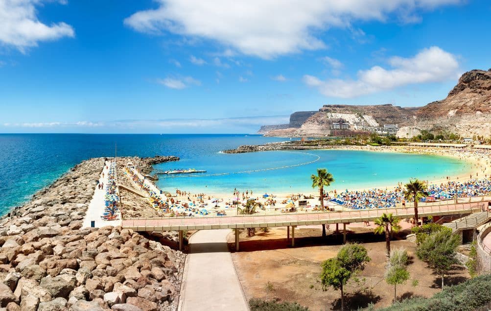 Vista dall'alto di una spiaggia affollata in una cala con acqua turchese, circondata da scogliere e un pontile roccioso sotto un cielo blu.