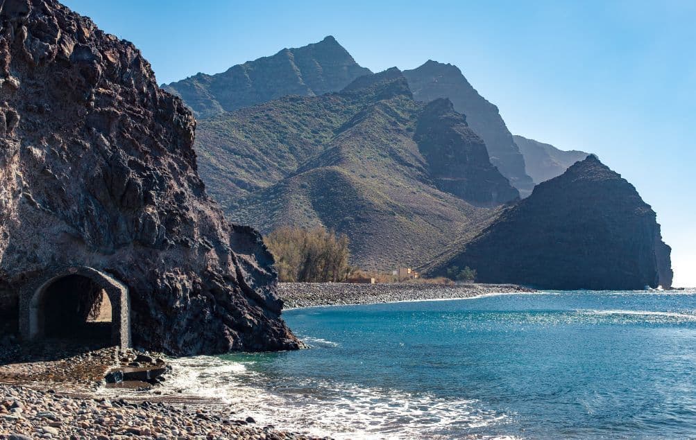 Un tunnel di pietra scavato in una scogliera si apre su una spiaggia di ciottoli accanto al mare blu, con grandi montagne sullo sfondo.