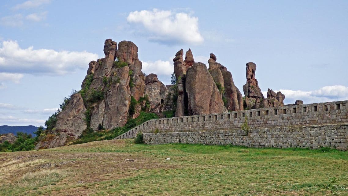 Una fortezza di pietra costruita in imponenti formazioni rocciose rossastre su una collina erbosa sotto un cielo parzialmente nuvoloso.