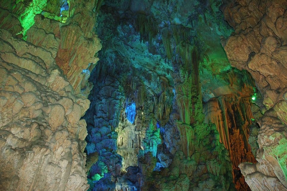 Una grande grotta con numerose stalattiti e formazioni rocciose illuminate da luci verdi e blu.