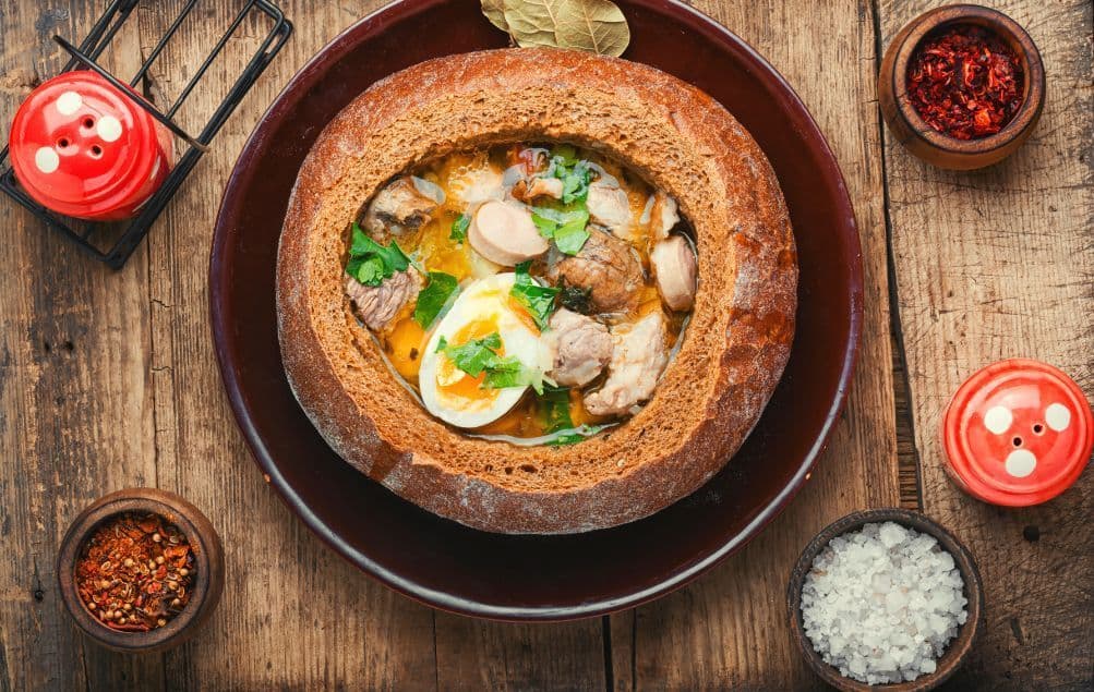 Eine Draufsicht auf eine Suppe mit Fleisch, Wurst und einem hartgekochten Ei, serviert in einem ausgehöhlten Brotleib auf einem Holztisch.