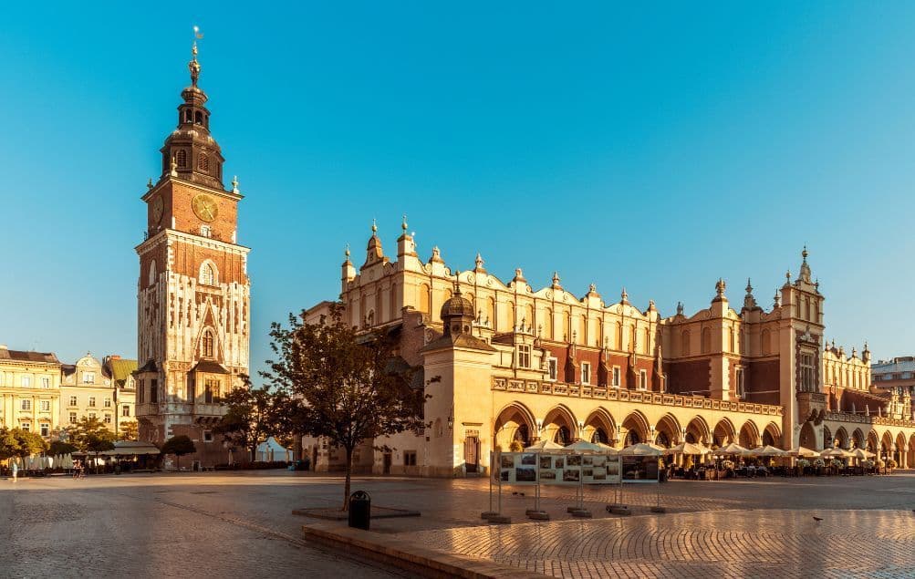 Ein historischer Glockenturm und ein verziertes Gebäude mit Arkaden stehen auf einem großen Kopfsteinpflasterplatz unter einem klaren blauen Himmel.
