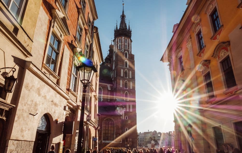 Ein historischer Backsteinkirchturm, von einer belebten Straße aus gesehen, mit einem hellen Sonnenstern und Linsenreflexion vor klarem blauem Himmel.