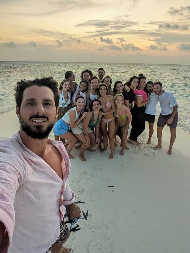 A man takes a selfie holding a drone with a WeRoad group trip posing on a white sand beach at sunset.