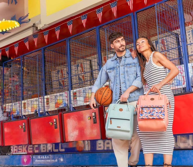 Ein Mann und eine Frau posieren mit Rucksäcken vor einem Jahrmarkt-Basketballspiel, das mit amerikanischen Flaggenwimpeln geschmückt ist.